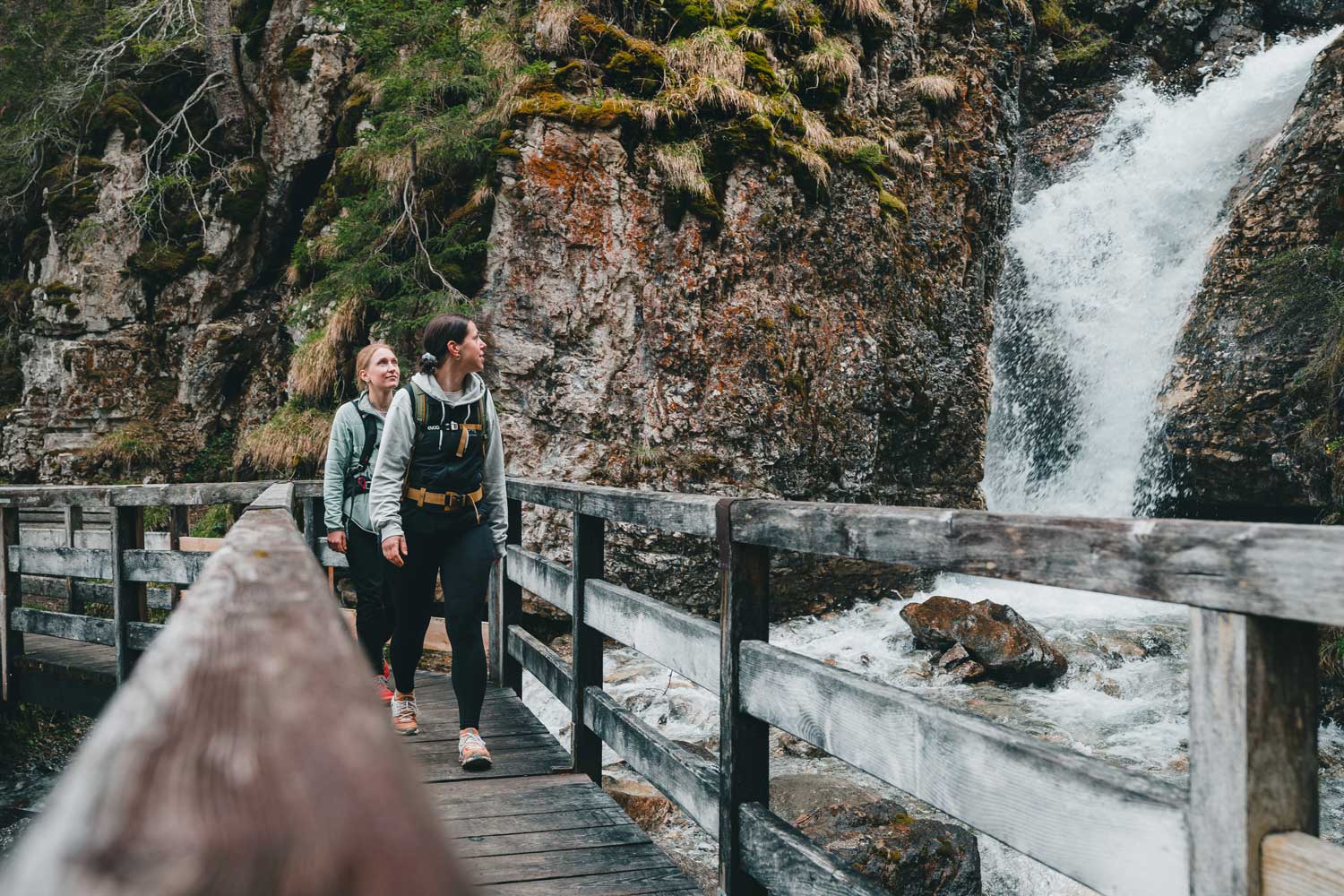 Zwei Wanderinnen gehen über eine Brücke neben einem Wasserfall